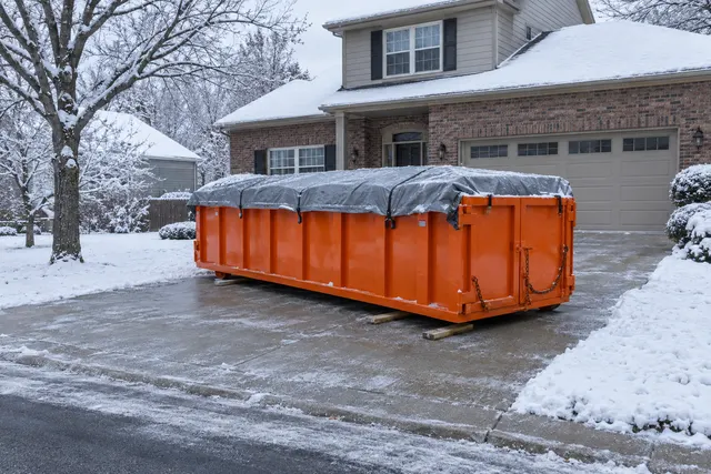 Orange roll off dumpster with a gray tarp strapped over the load, sitting on boards on a shoveled driveway in front of a brick Kansas City home after a snowfall